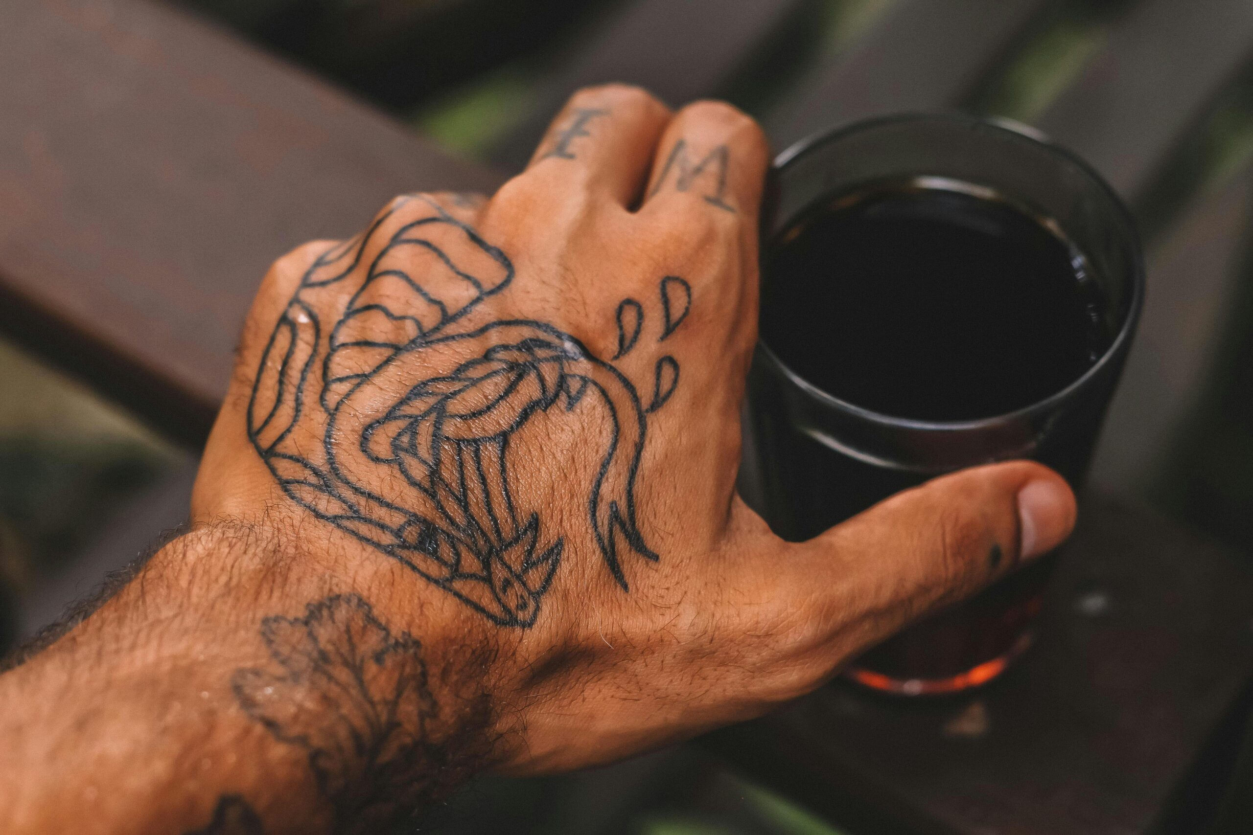 A close-up shot of a tattooed hand holding a glass of black coffee outdoors.